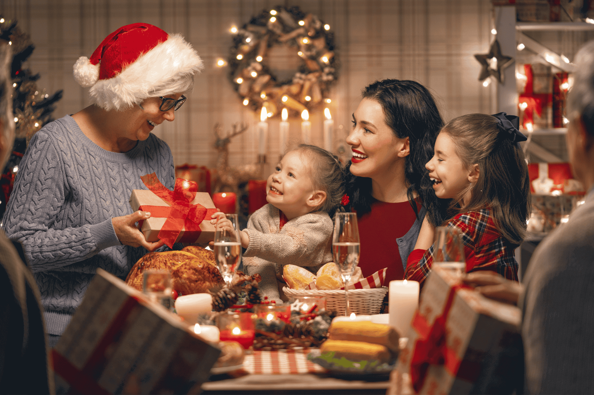 Mehrere Generationen einer Familie sitzen an einem festlich gedeckten Weihnachtstisch, lachen und überreichen ein Geschenk.