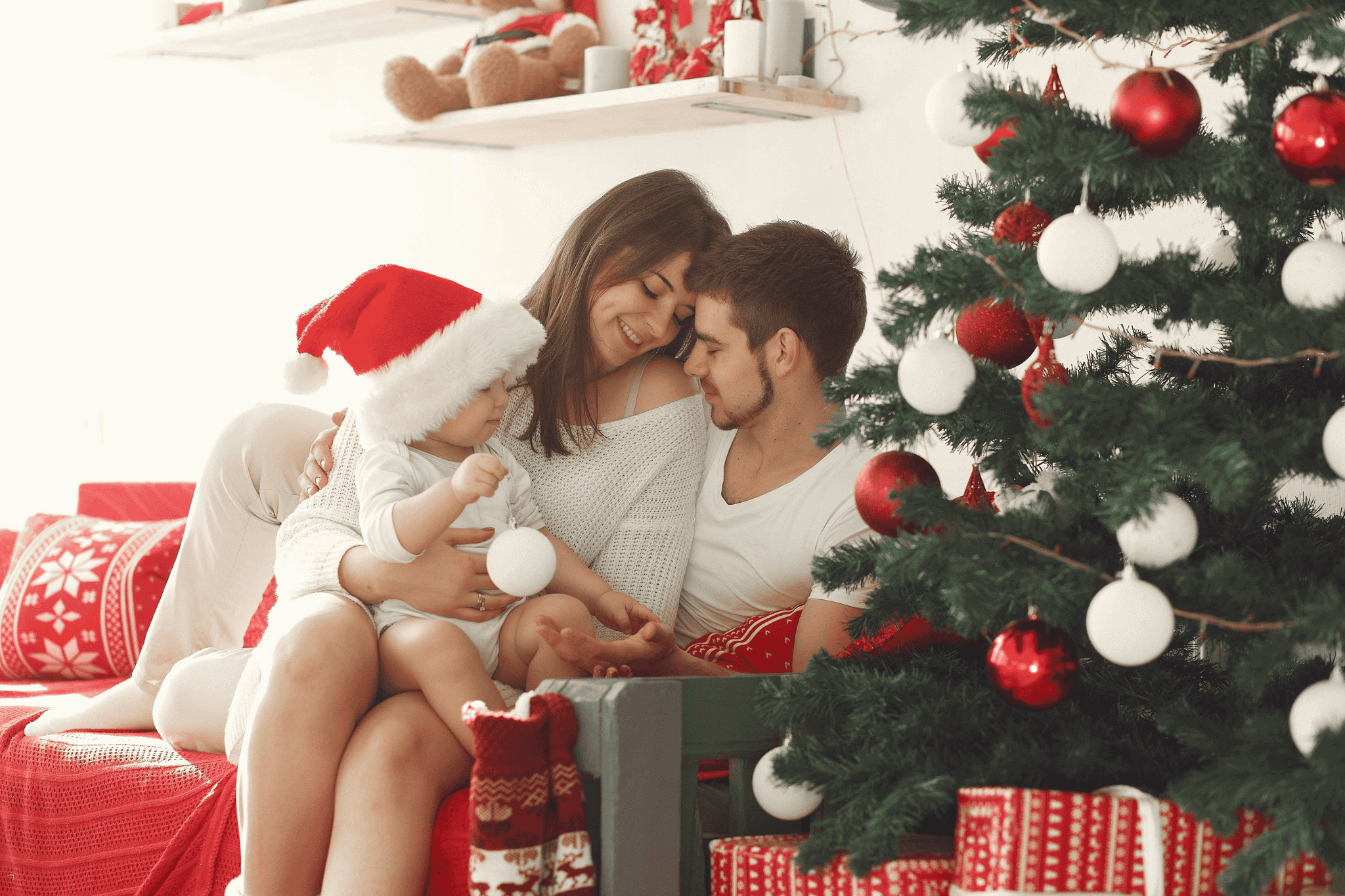 Eltern halten ihr Baby vor einem Weihnachtsbaum, während im Hintergrund ein Kamin brennt und eine warme, festliche Atmosphäre entsteht.