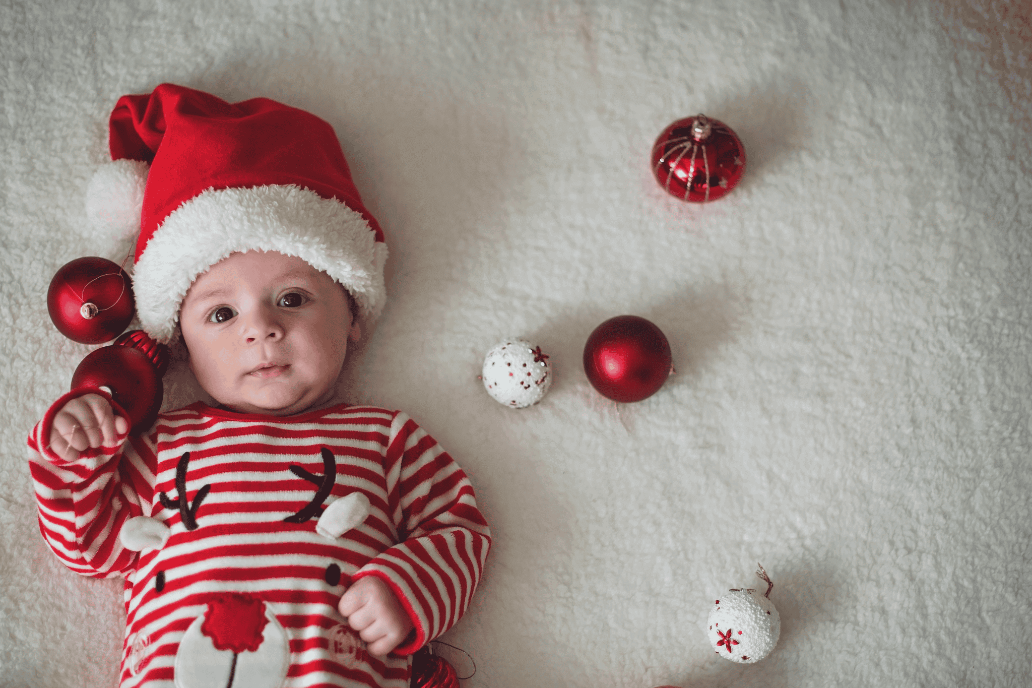 Ein Baby liegt auf einer weißen Decke, trägt eine rote Weihnachtsmütze und einen gestreiften Weihnachtspyjama, umgeben von Weihnachtsbaumkugeln.