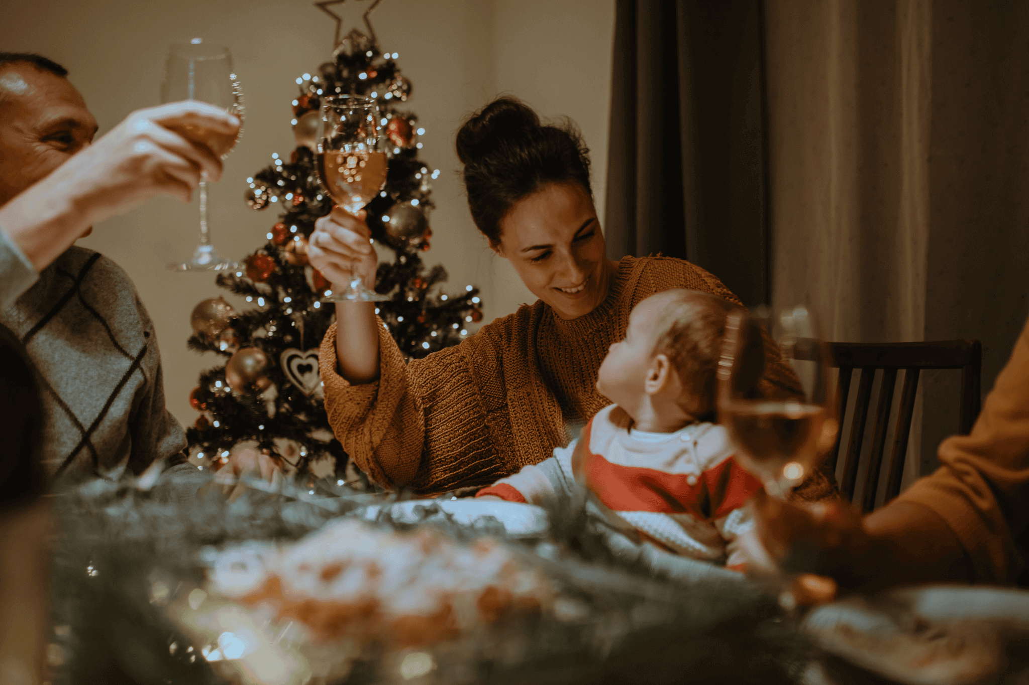 Eine Familie sitzt am festlich gedeckten Tisch, stößt mit Gläsern an und feiert Weihnachten, während ein Baby auf dem Schoß eines Erwachsenen sitzt