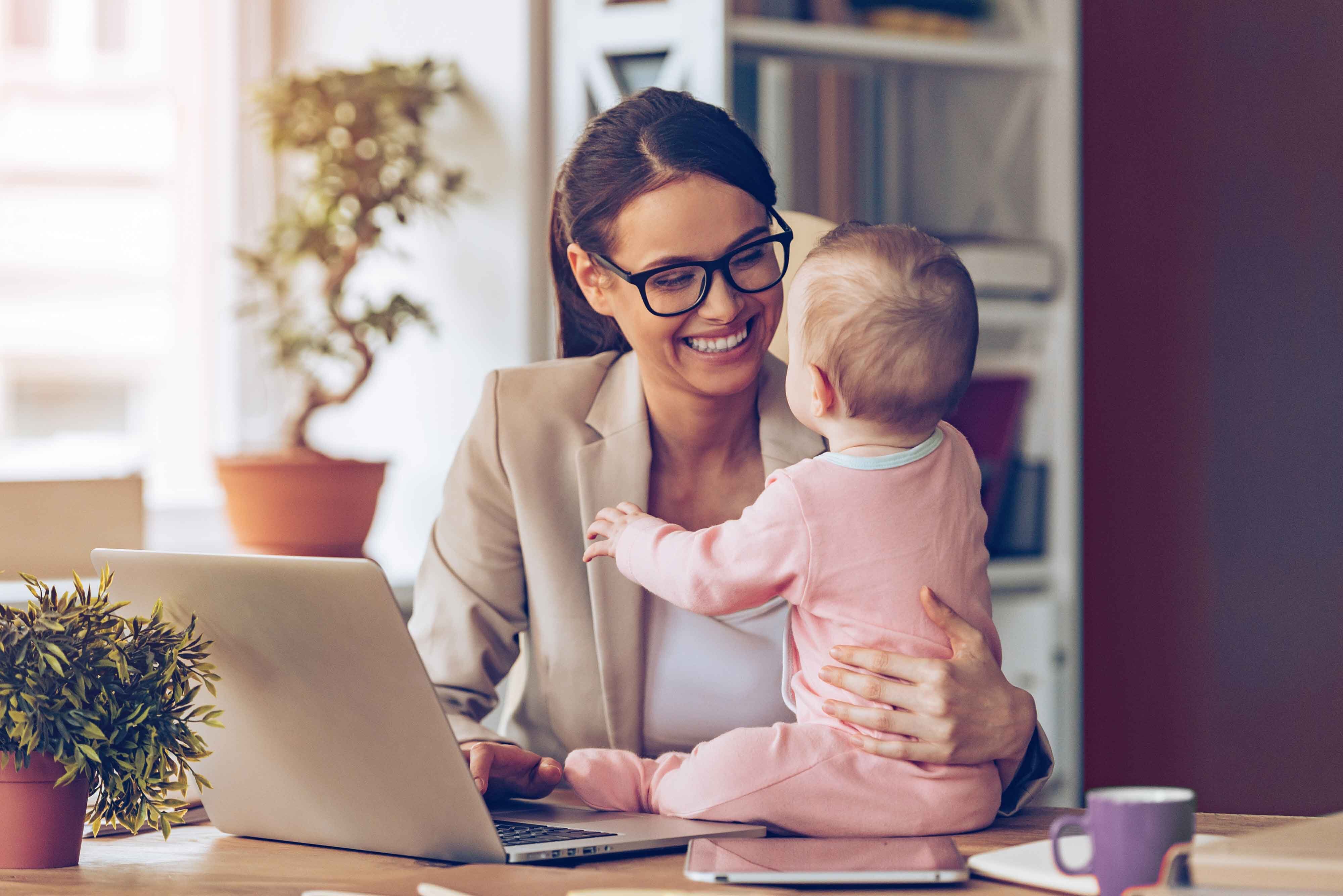 Eine Frau in Businesskleidung sitzt mit ihrem Baby am Schreibtisch