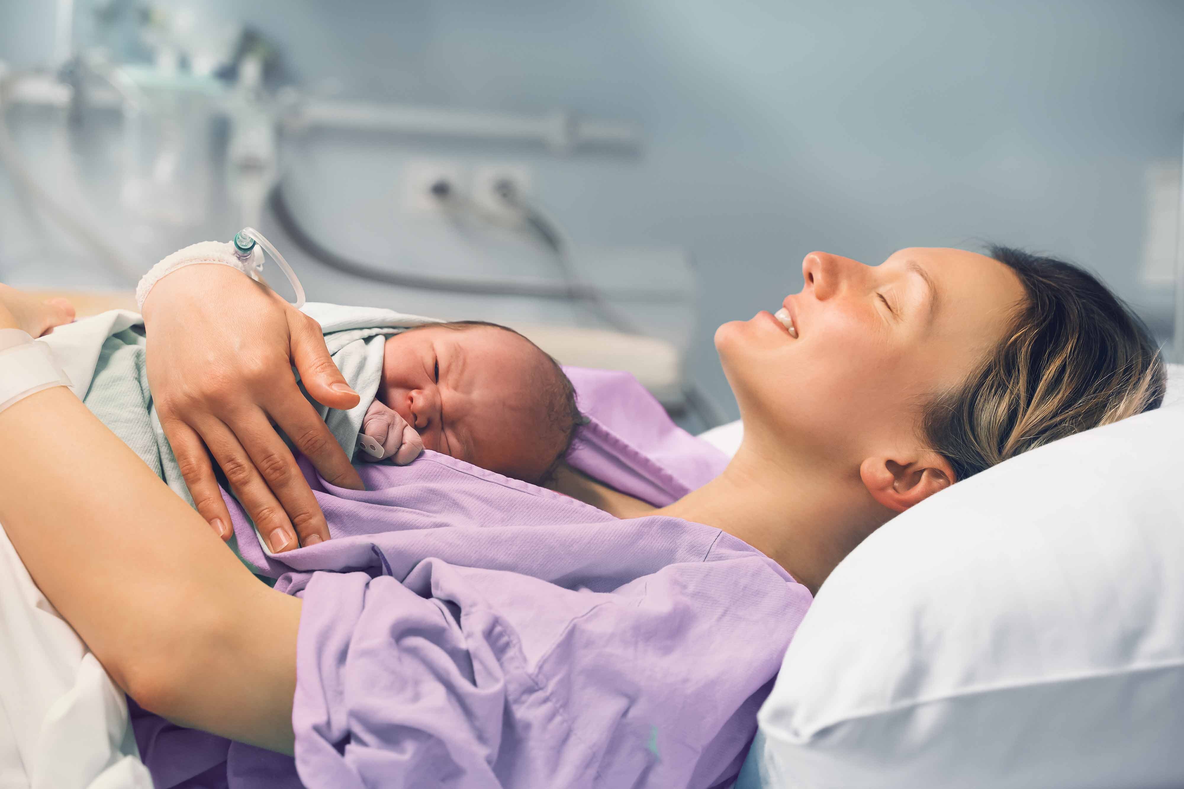 Mutter liegt mit schreiendem Saeugling im Bett einer Klinik